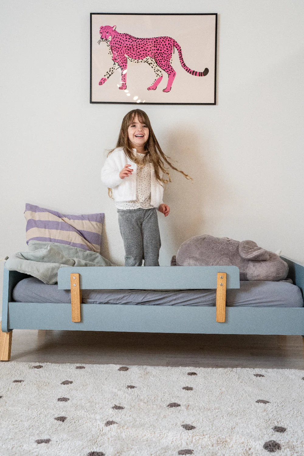 Child standing on a bed with a colorful painting on the wall.