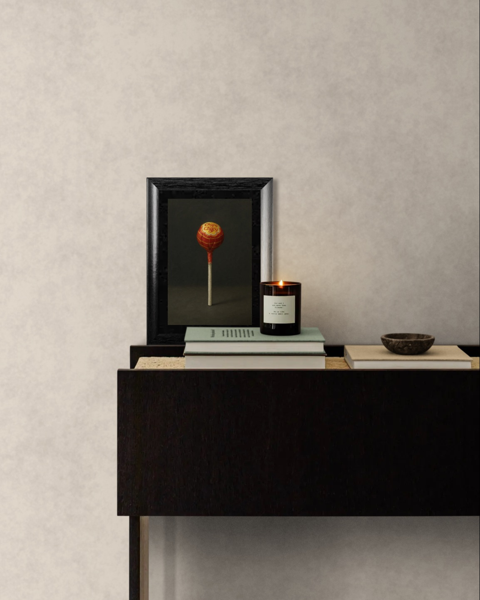 Framed artwork of a lollipop, candle, and books on a dark wooden console table against a light gray wall.